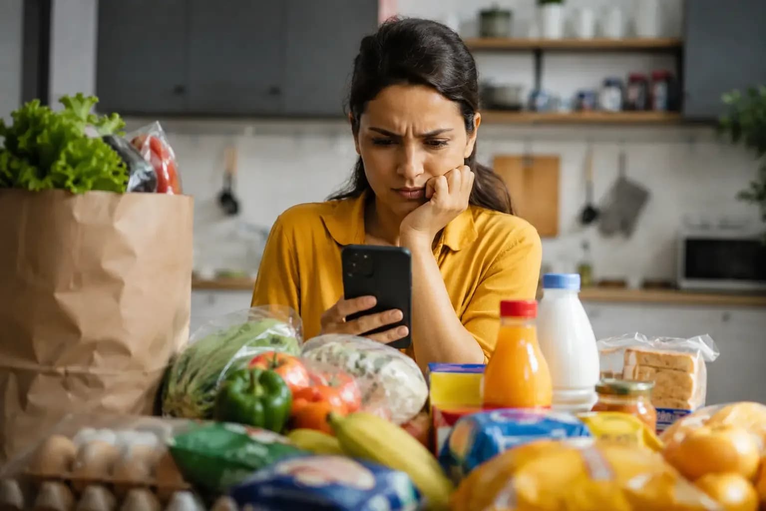 Woman reviewing a grocery order on her smartphone with groceries spread across the kitchen table, illustrating how quick commerce may influence household grocery spending in India.